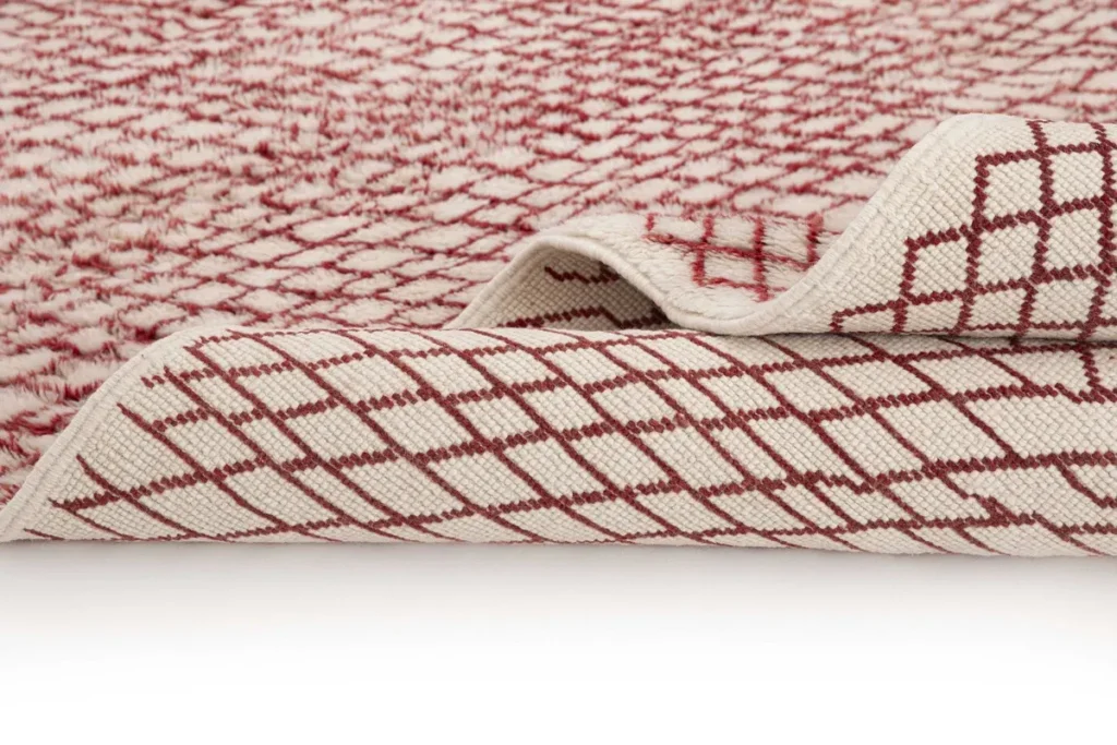 This luxurious, hand-knotted rug showcases a striking red-and-white pattern. Its close-up view highlights a grid-like design on both sides, with one edge artfully folded to reveal the intricate craftsmanship of the underside.