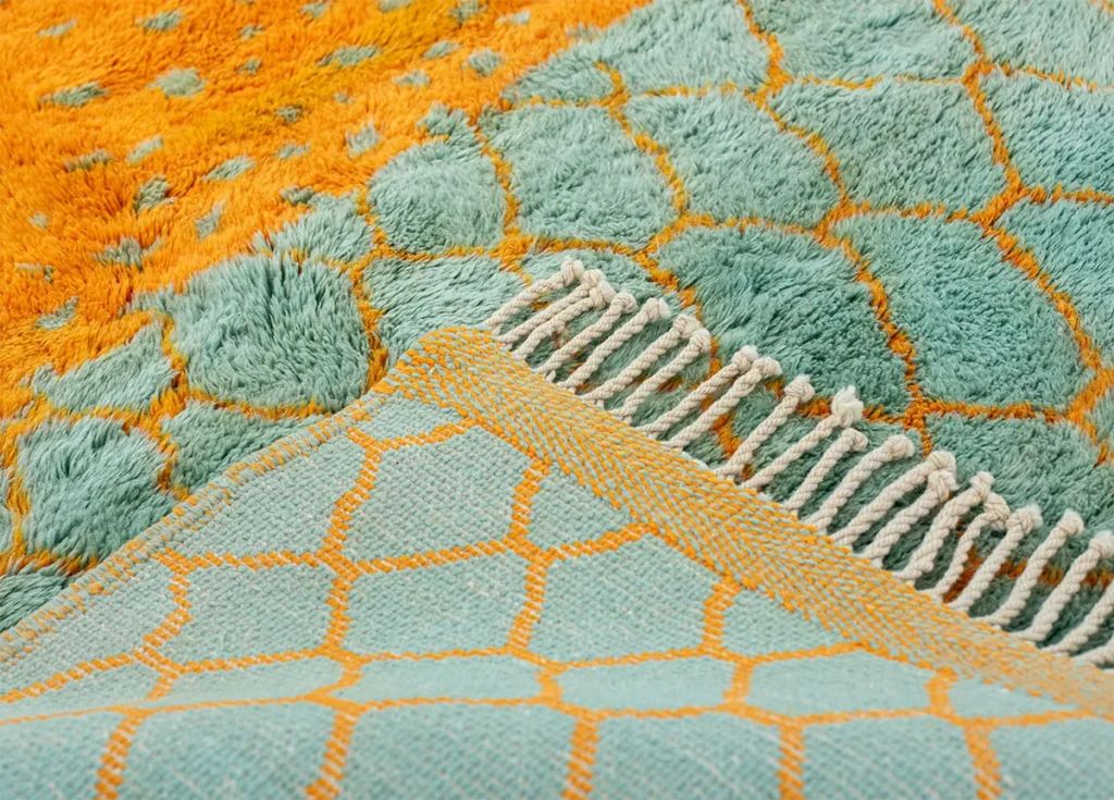 Close-up of a hand-knotted fabric with a geometric pattern in teal and orange hues. The fabric features a fringe and thick pile, showcasing an intricate design of hexagons and diamond shapes.