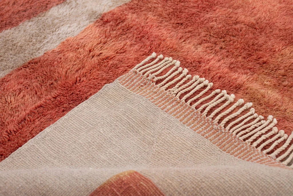 This close-up showcases an authentic Moroccan rug with an intricate hand-knotted design in shades of orange and fringed edges, partially folded to reveal a contrasting beige underside.