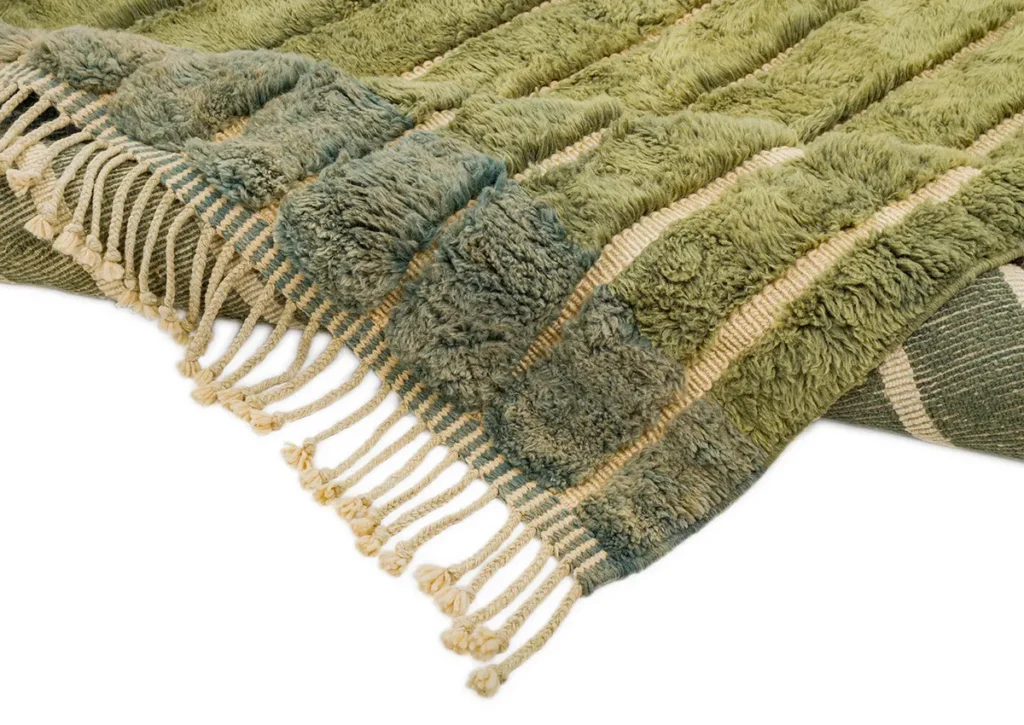 A close-up of a green and beige striped luxury rug with fringed edges, featuring textured, fluffy sections alternating with flat fabric.