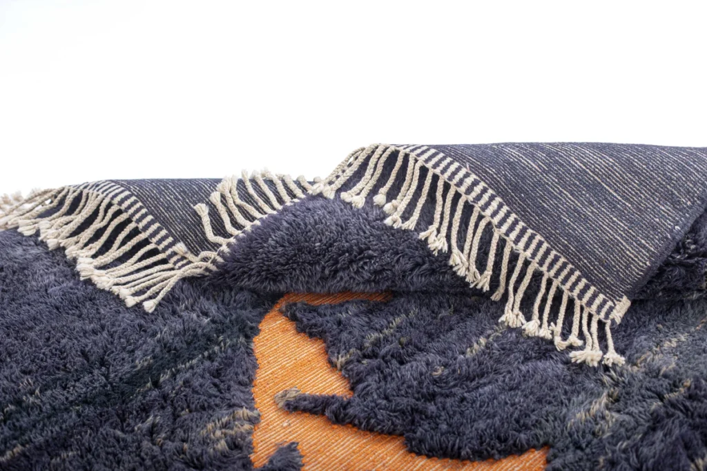 Close-up of a vibrant folded blue and orange woven rug with intricate fringe detailing against a pristine white backdrop.