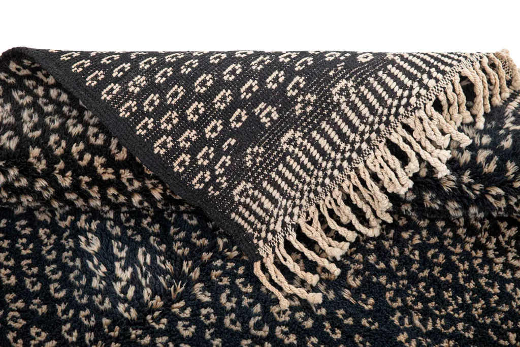 A hand-knotted black and beige woven fabric with intricate patterns and fringed edges, skillfully folded to reveal both sides.