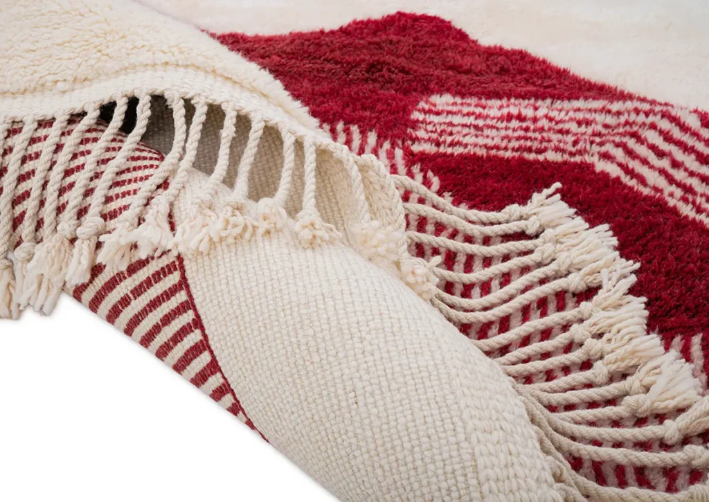 A close-up of two elegant, hand-knotted rugs shows a cream textile with red stripes alongside a vivid red piece adorned with a geometric pattern. Luxurious and intricate, these beautifully crafted pieces bring sophistication to any space.