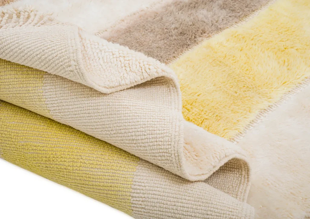 Experience the elegance of luxury rugs with a close-up of a folded plush rug, showcasing cream, beige, and yellow stripes. Notice both the soft and textured sides of this exquisite piece, perfect for adding sophistication to any space.