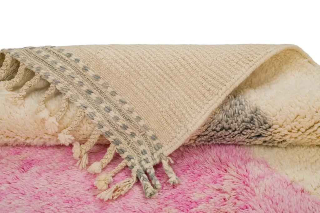 A folded woven rug with tassels on one edge is elegantly displayed on a plush pink and white surface, adding a touch of sophistication.