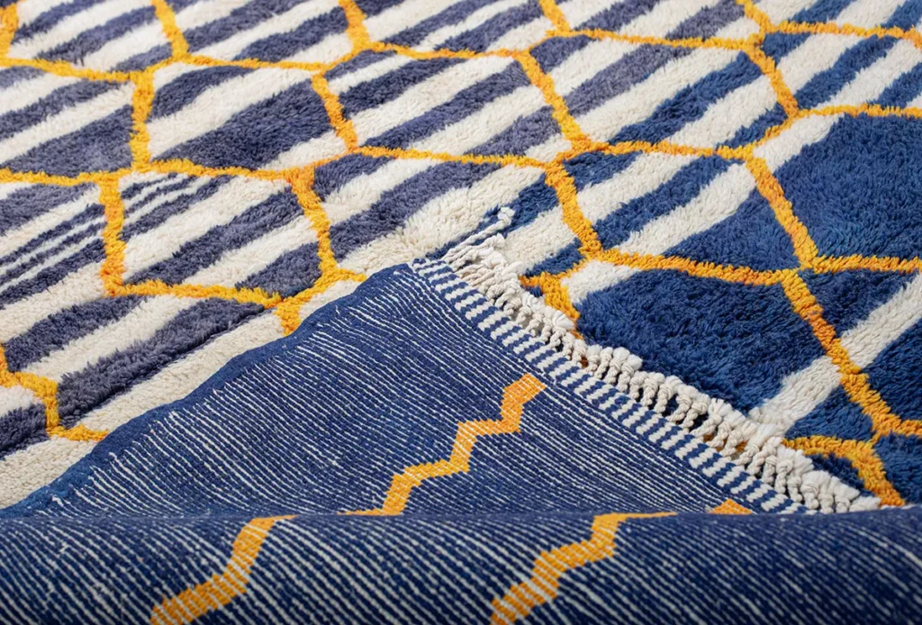 This luxury rug showcases a stunning blue and yellow pattern with a folded corner revealing a striped underside. Its intricate design features geometric shapes and lines, perfectly embodying the artistry of hand-knotted rugs.