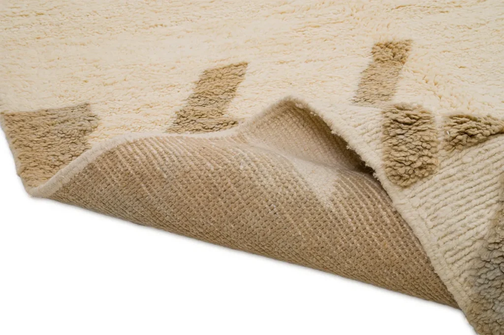 A luxurious beige textured rug with brown geometric patterns is partially folded to reveal its underside, showcasing the intricate craftsmanship typical of a hand-knotted Moroccan rug.