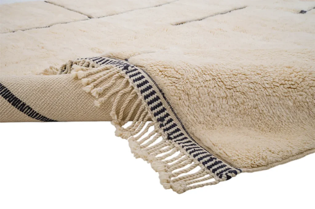 Close-up of an artisanal rug with beige texture and black striped fringe edges. The hand-knotted rug is folded at one corner, revealing its woven underside.
