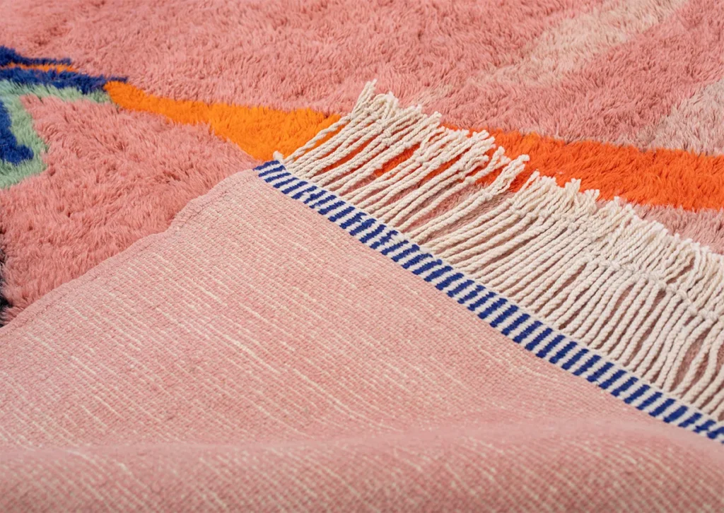 Close-up of a fringed, pink fabric with a blue-striped border gracefully draped over a plush Moroccan rug showcasing pink, orange, and multicolored geometric patterns.