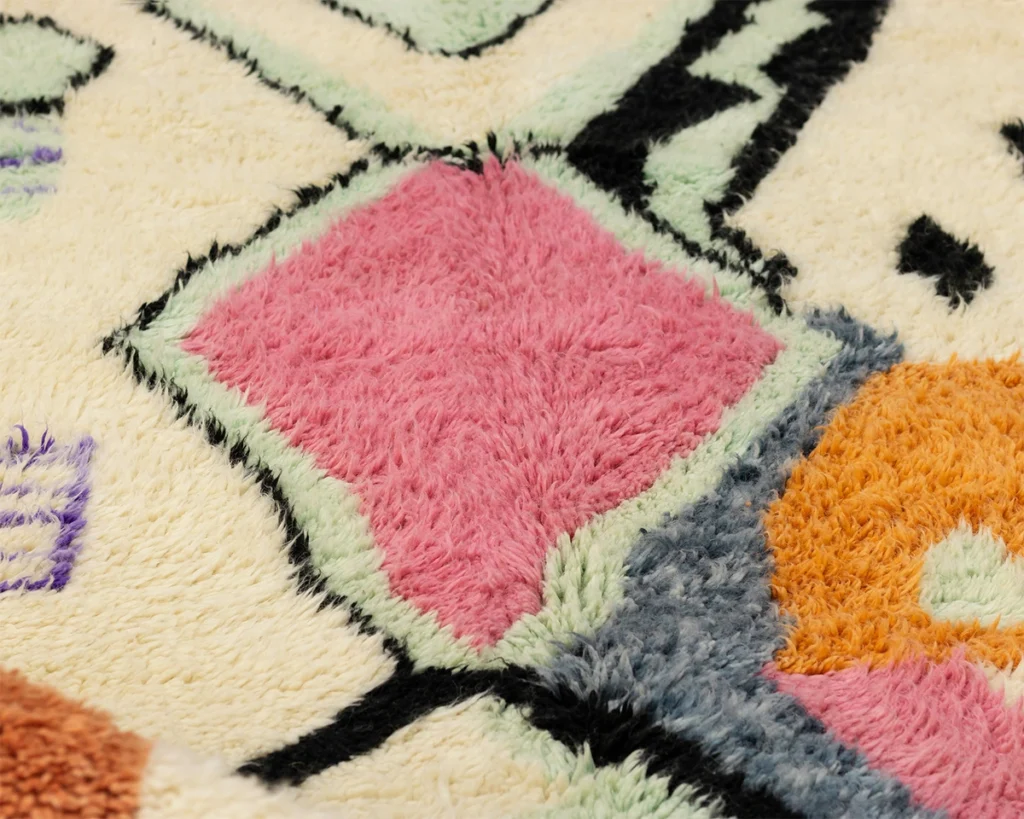 This close-up showcases an artisanal rug with textured geometric patterns in pink, orange, green, and black on a cream background. Hand-knotted details enhance its beauty, offering a sophisticated touch to any space.
