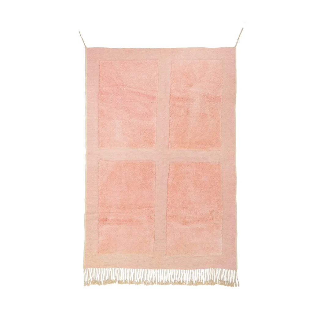 A pink rectangular rug with four eye-catching quadrants hangs vertically. Adorned with fringed ends, its elegantly suspended by two cords, adding a touch of eclectic style to any space.