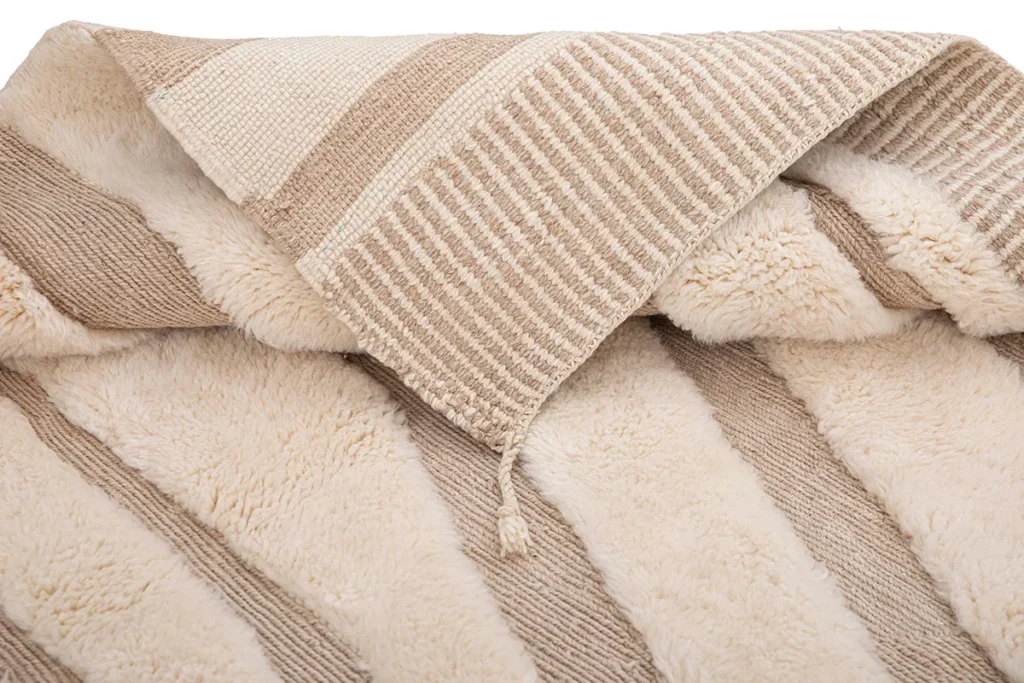 A close-up of a woven texture with plush lining, featuring beige and cream stripes. A tassel adds an elegant touch to this piece, reminiscent of luxury rugs.