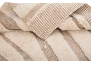 A close-up of a woven texture with plush lining, featuring beige and cream stripes. A tassel adds an elegant touch to this piece, reminiscent of luxury rugs.