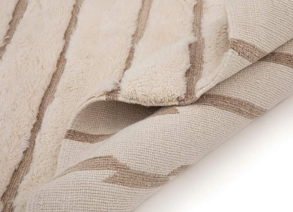 A close-up of a beige and brown striped luxury rug with a corner flipped over, revealing its woven underside.