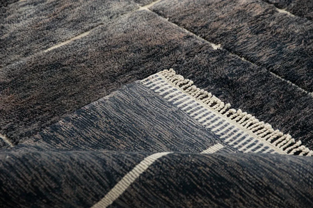 Close-up of an artisanal rug showcasing a textured, dark color with a woven pattern and fringe detail. This hand-knotted piece features alternating stripes and varied textures, adding a touch of elegance to any room.