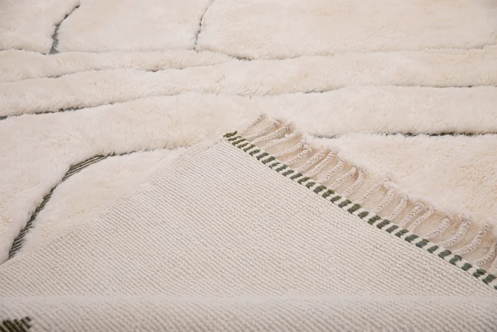 This elegant close-up showcases the corner of a cream-colored luxury rug on a matching carpet. The hand-knotted rug features a woven texture with green-striped tassels, adding an exquisite touch to any space.