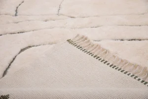 This elegant close-up showcases the corner of a cream-colored luxury rug on a matching carpet. The hand-knotted rug features a woven texture with green-striped tassels, adding an exquisite touch to any space.