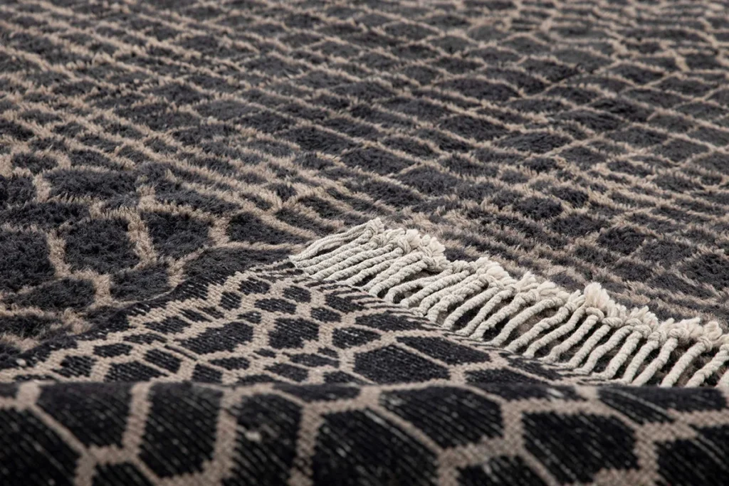 This luxurious close-up showcases a textured fabric with a black and beige geometric pattern, complemented by elegant fringe trim, reminiscent of hand-knotted rugs.