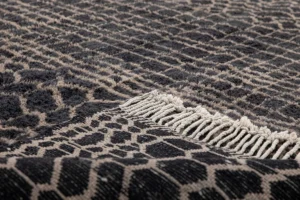 This luxurious close-up showcases a textured fabric with a black and beige geometric pattern, complemented by elegant fringe trim, reminiscent of hand-knotted rugs.