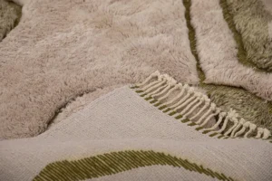 This luxurious Moroccan rug features a close-up of its beige and green woven fabric adorned with tassels. The hand-knotted design showcases intricate patterns and a soft, fluffy texture, exuding elegance and craftsmanship.