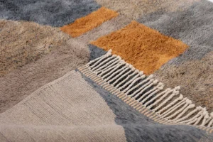 This close-up showcases an authentic Moroccan rug with fringed edges. The design features abstract geometric shapes in shades of gray, orange, and beige, reflecting artisanal craftsmanship and tradition.