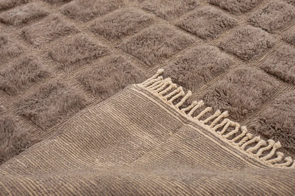 Close-up of a textured beige luxury rug with a checkered pattern and fringed edges, exuding an air of elegance.