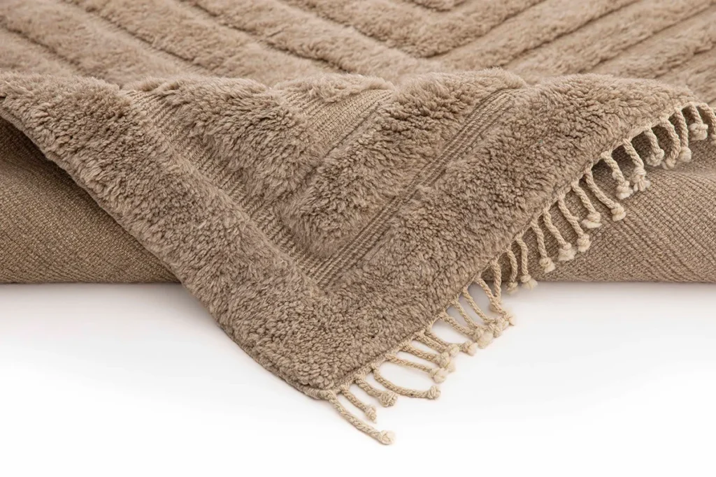 A close-up of a luxurious hand knotted rug in rich brown, showcasing its intricate texture and fringe detail, elegantly laid on a pristine white surface.