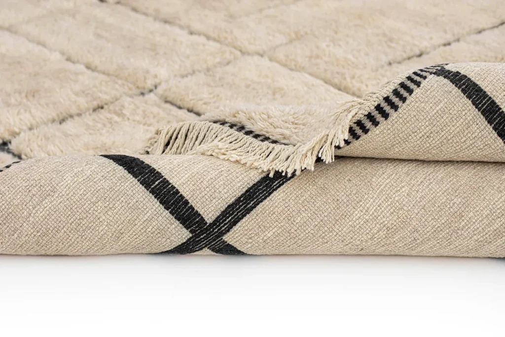 A luxurious folded beige rug, hand-knotted with black geometric lines and fringed edges, sits elegantly on a white background.