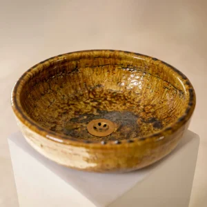 A round, rustic ceramic sink with a brown and amber glaze sits on a white pedestal against a neutral background.
