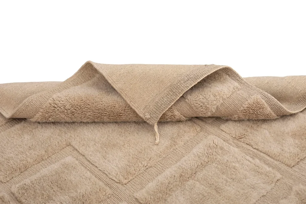 A tan luxury rug with a diamond-patterned surface is elegantly folded to showcase its textured underside.