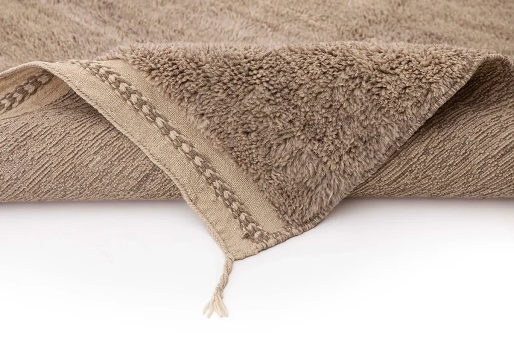 A close-up of a luxury rug with a textured surface and a decorative fringe on the edge, resting on a white background. The intricate details and vibrant patterns highlight exquisite craftsmanship.