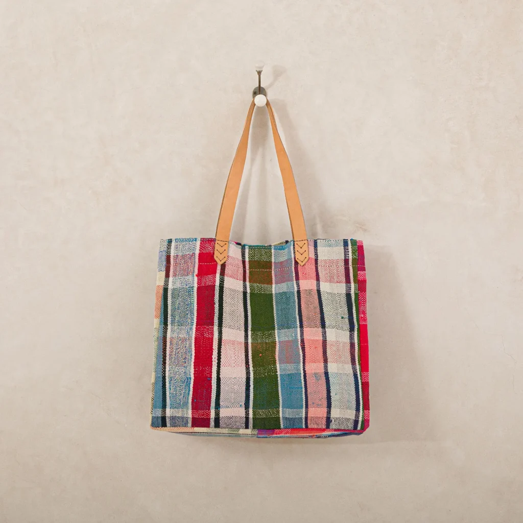 A colorful plaid fabric tote bag with tan leather handles hangs on a white wall hook.