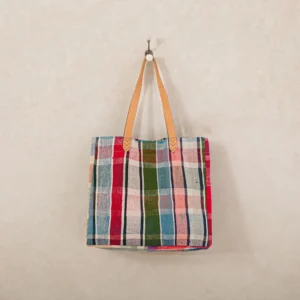 A colorful plaid fabric tote bag with tan leather handles hangs on a white wall hook.