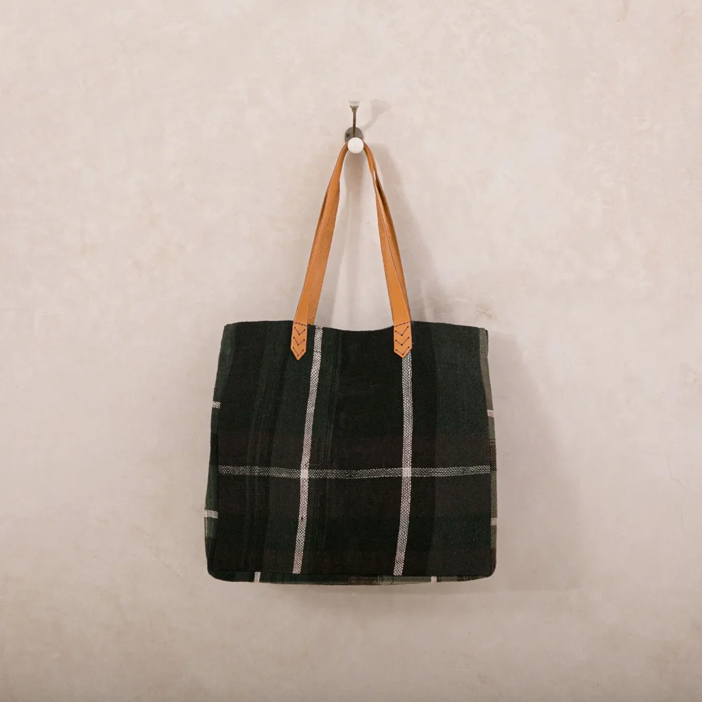 A black-and-white plaid tote bag with tan leather handles hangs on a wall hook.