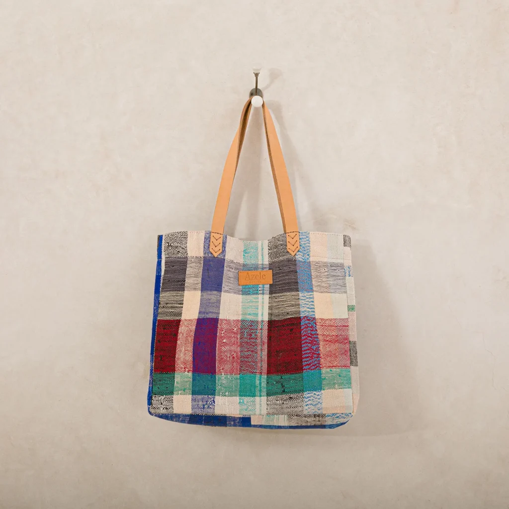 A multicolored patchwork tote bag with tan handles hangs on a wall hook.