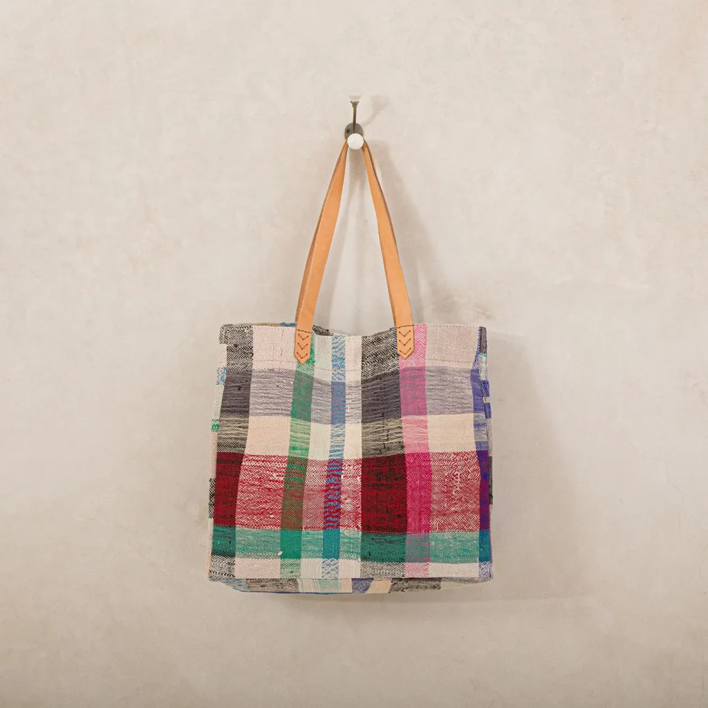 A patchwork tote bag with colorful squares and light brown leather handles hangs on a wall hook against a beige background.