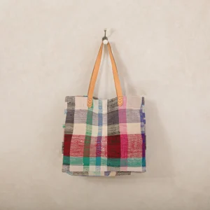 A patchwork tote bag with colorful squares and light brown leather handles hangs on a wall hook against a beige background.