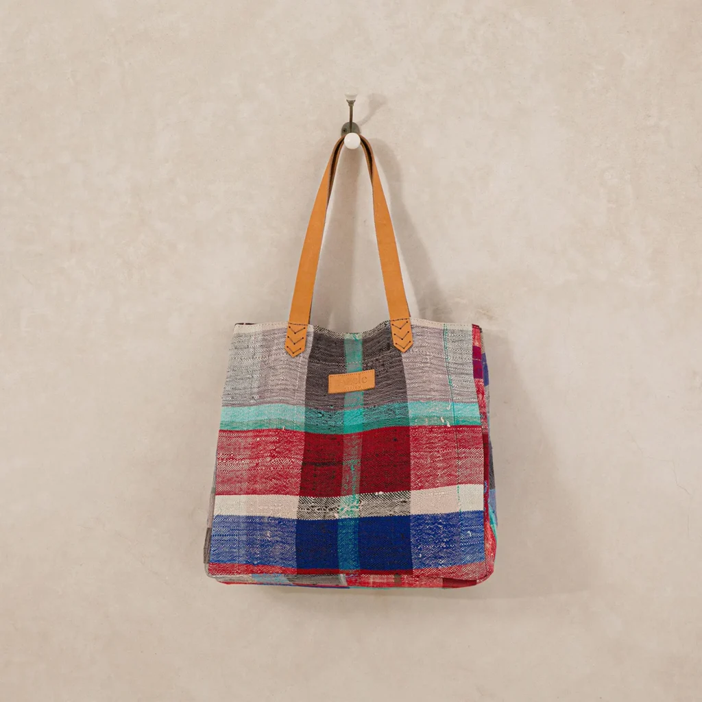 Colorful plaid tote bag with tan leather straps hanging on a wall hook.