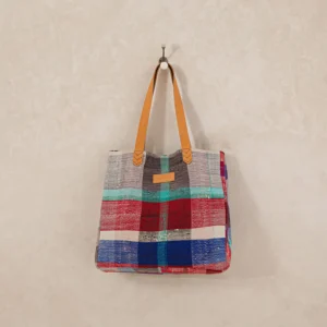 Colorful plaid tote bag with tan leather straps hanging on a wall hook.
