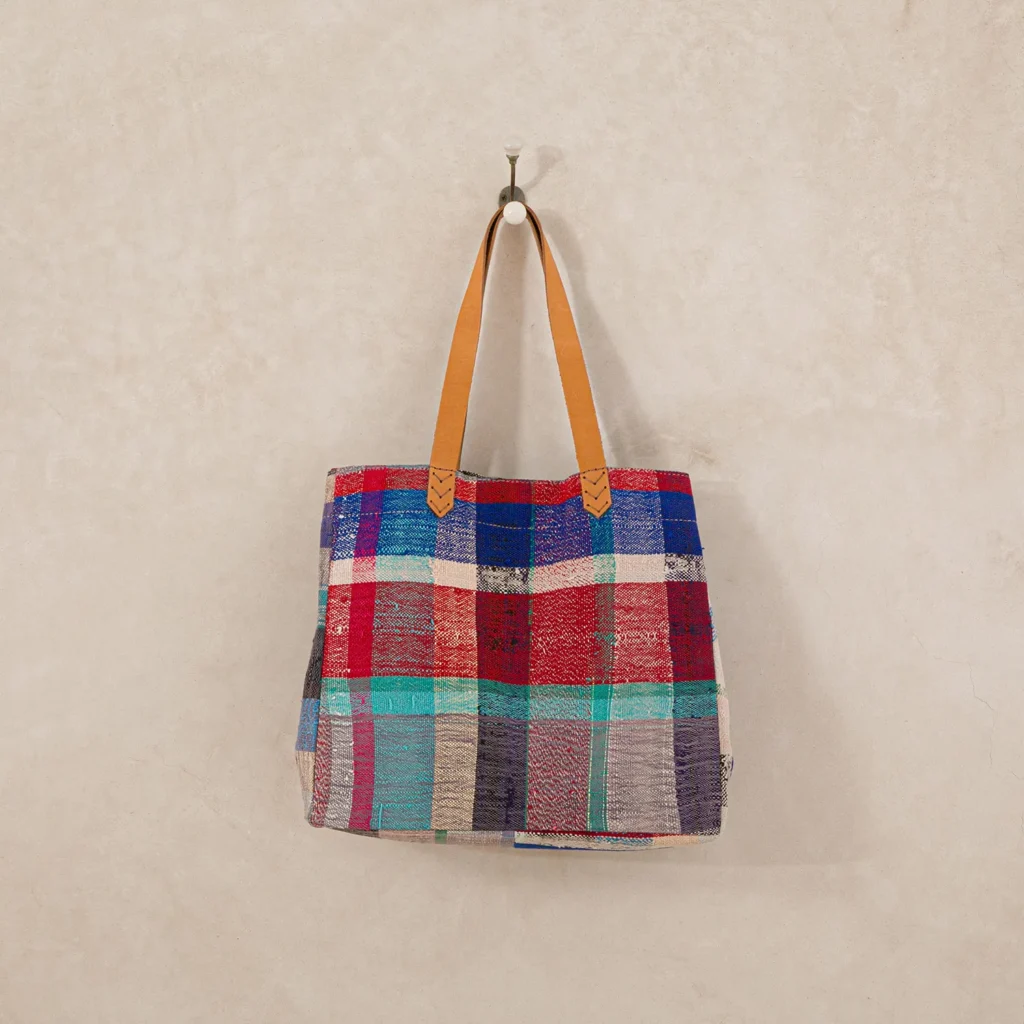 A multicolored checkered fabric tote bag with tan leather handles hangs on a wall hook.