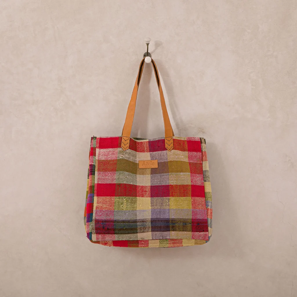 A multicolored plaid fabric tote bag with brown leather handles hangs on a wall hook.