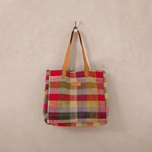 A multicolored plaid fabric tote bag with brown leather handles hangs on a wall hook.
