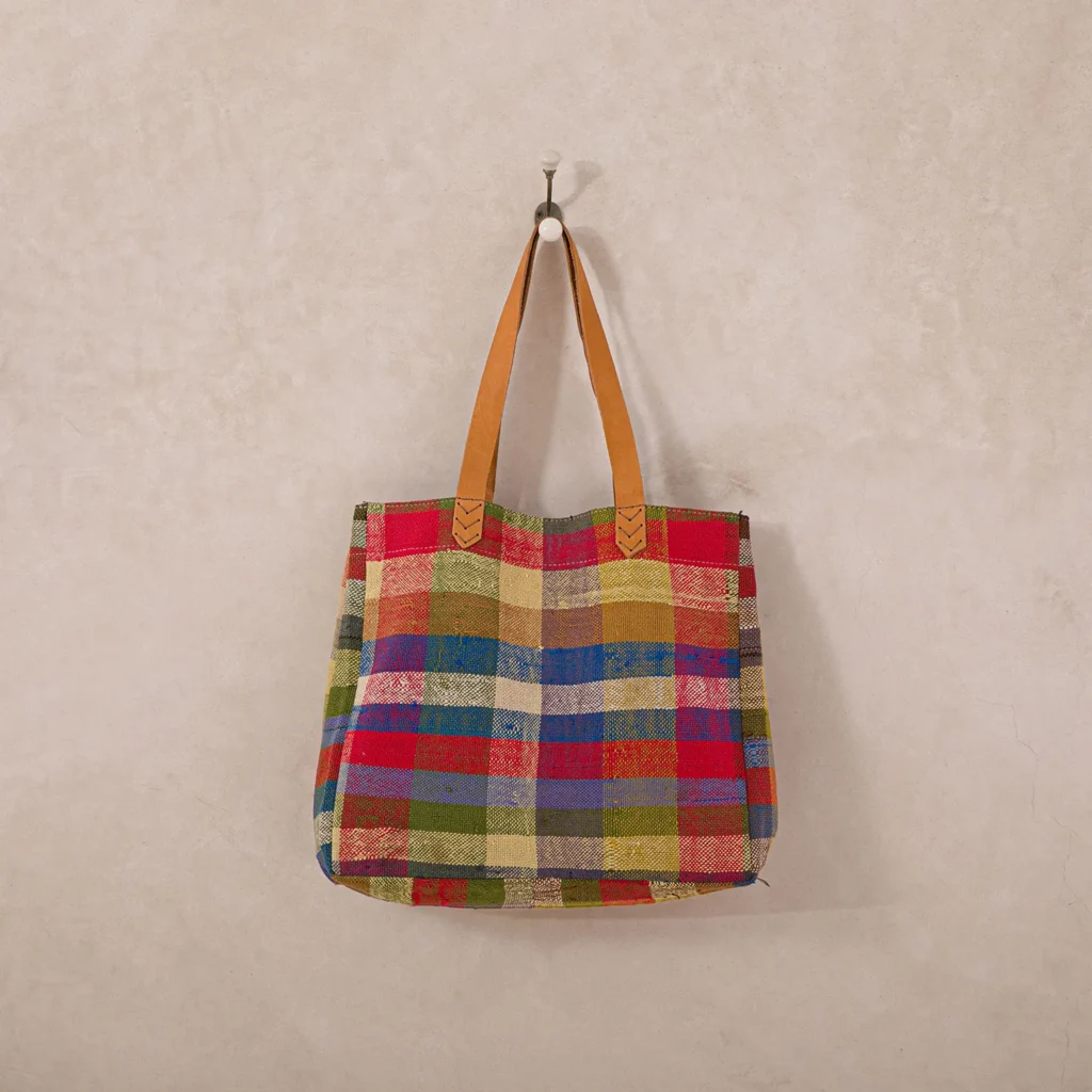 A colorful plaid tote bag with brown handles hangs on a wall hook.