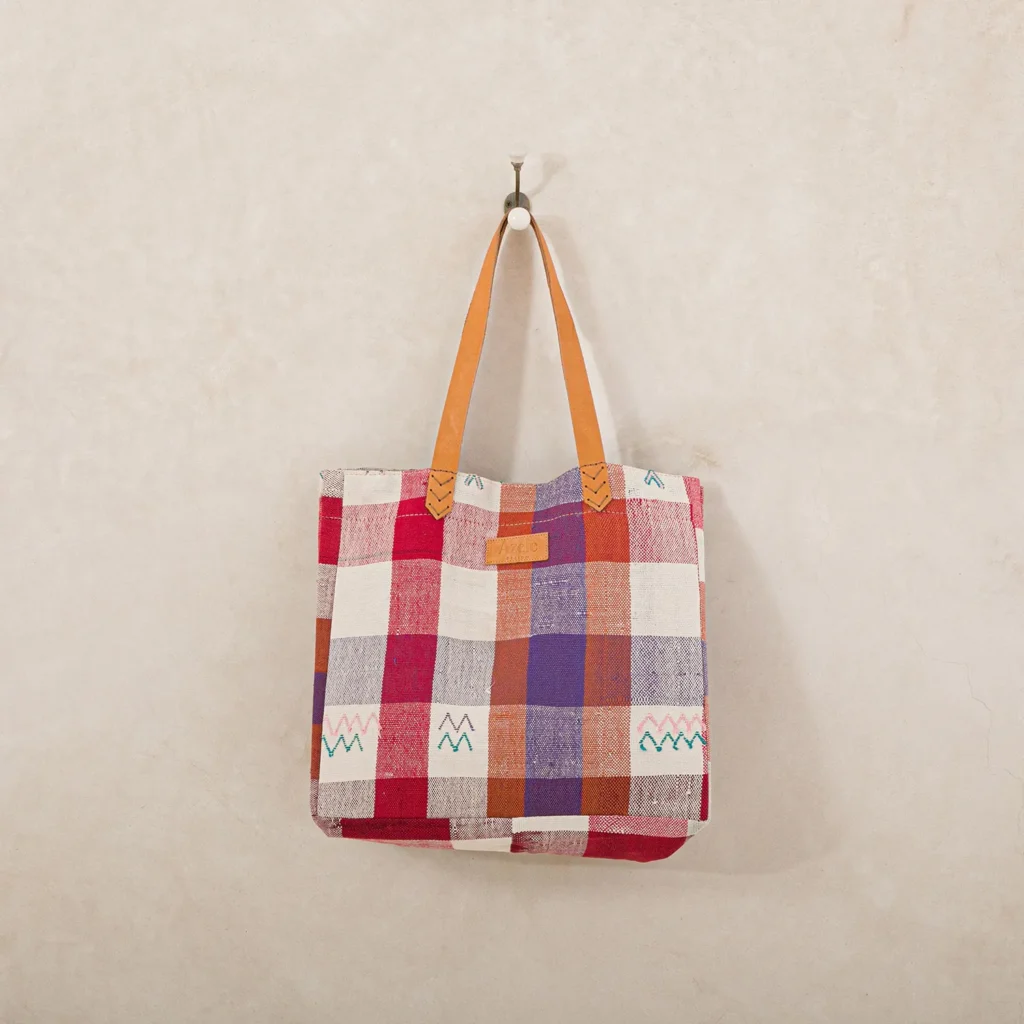A multicolored checkered tote bag with orange handles hangs on a hook against a plain wall.