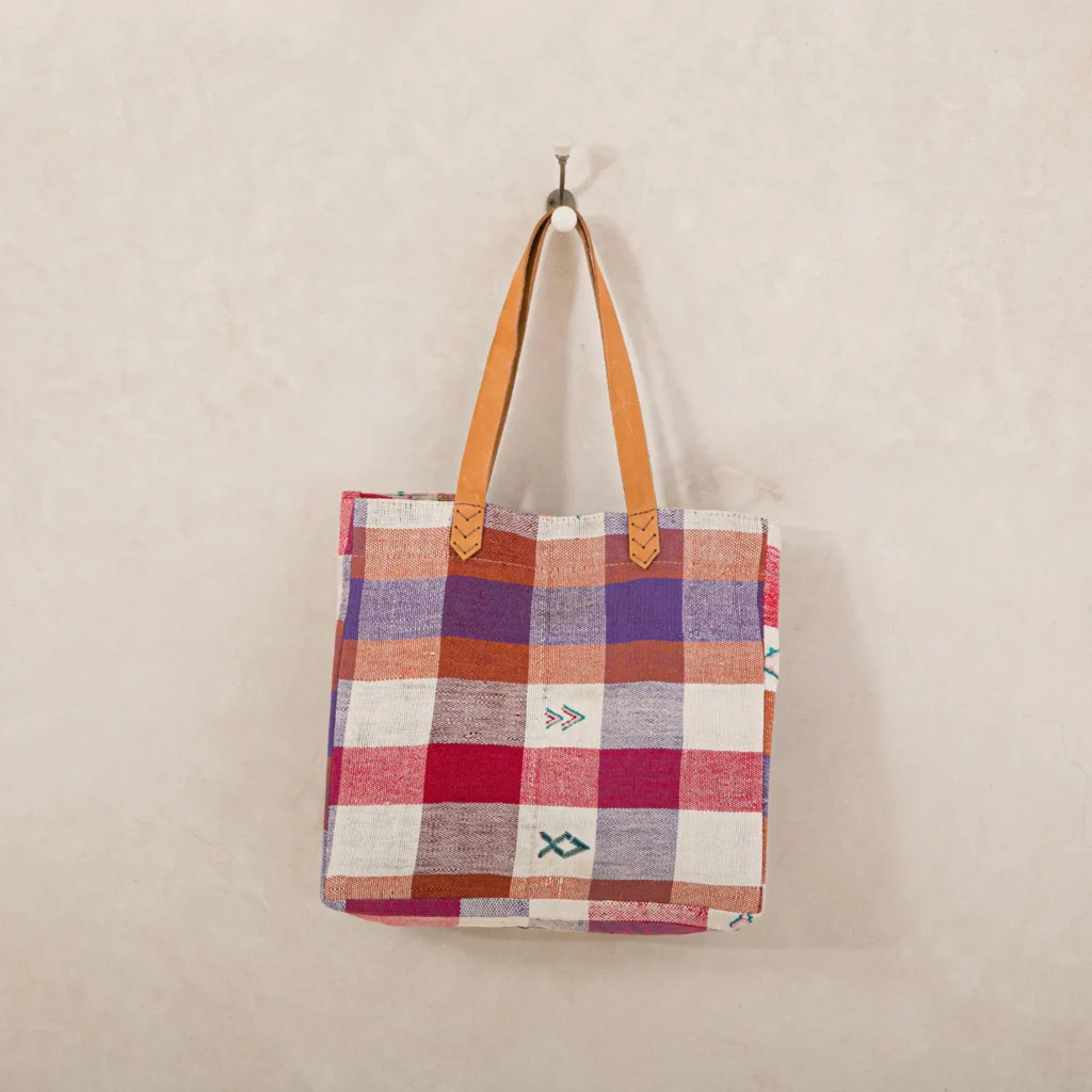 A checkered tote bag with leather handles hangs on a wall hook. The pattern features red, purple, and white squares, with small arrow-like designs.