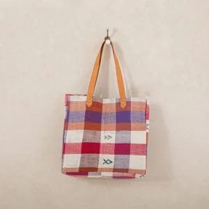 A checkered tote bag with leather handles hangs on a wall hook. The pattern features red, purple, and white squares, with small arrow-like designs.