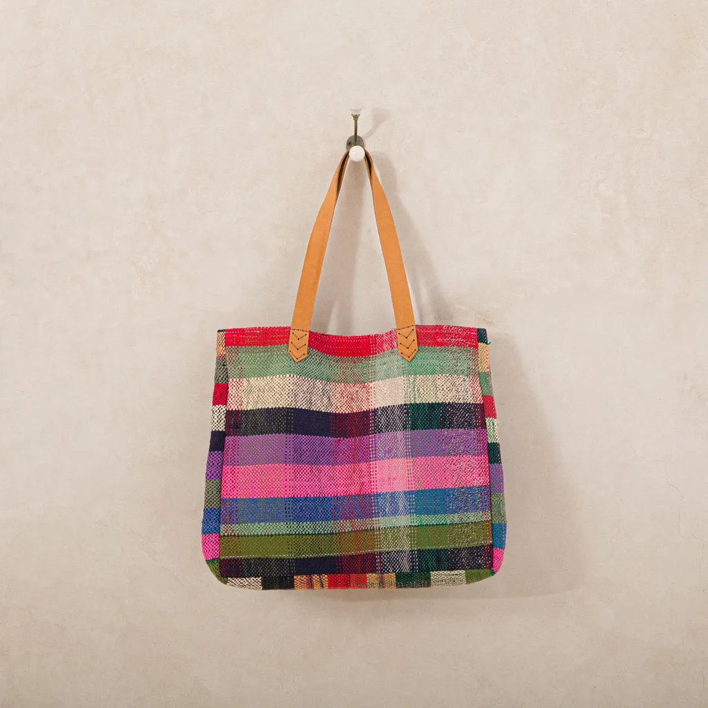 A multicolored woven tote bag with brown leather handles hangs on a hook against a plain wall.