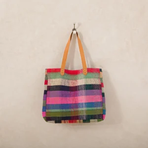 A multicolored woven tote bag with brown leather handles hangs on a hook against a plain wall.