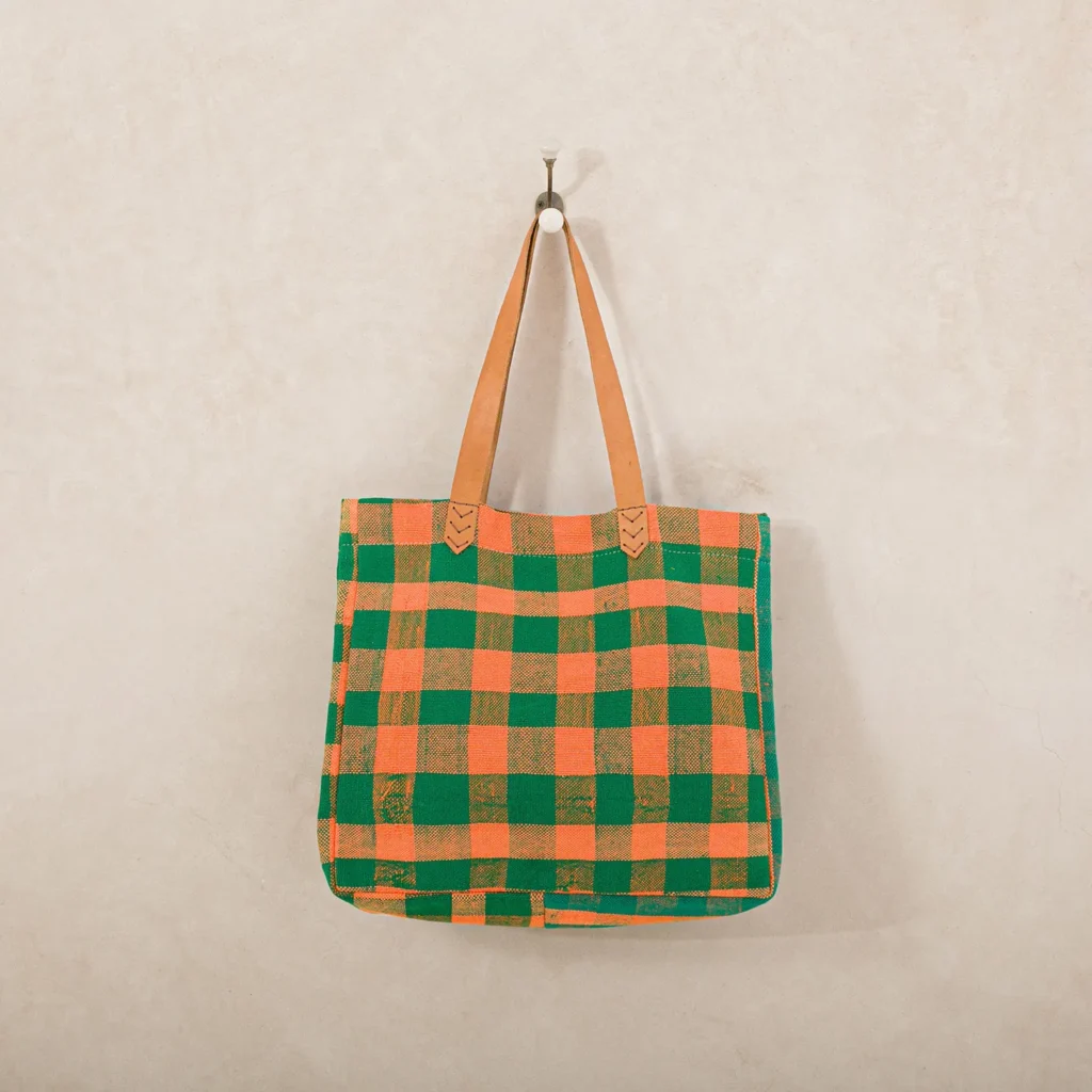 A checkered tote bag in green and orange with tan handles is hanging on a hook against a beige wall.
