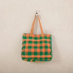 A checkered tote bag in green and orange with tan handles is hanging on a hook against a beige wall.
